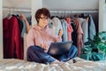 Young mixed race woman in eyeglasses with cup of coffee working on laptop while sitting on bed. Student's Apartment