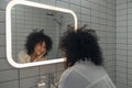 Young mixed race woman looking herself in the mirror with love. Smiling female with curly hair in a modern washroom. Royalty Free Stock Photo