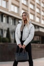 Young model of a beautiful woman with stylish black bag in a fashionable white jacket in jeans stands outdoors on an autumn day Royalty Free Stock Photo