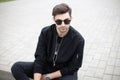 Young model man in fashionable black clothes in dark sunglasses rest sitting on a tile in the city on a bright summer day. Royalty Free Stock Photo