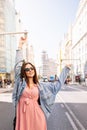 Young model smiling, posing and opening arms in an pink dress, denim jacket, boots and eye cat sunglasses standing at the famous