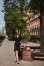 A young model woman in a black dress and sunglasses walking by the sidewalk and enjoying a sunny summer day Royalty Free Stock Photo