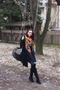 Young modern caucasian woman in black coat and colorful scarf outdoor pose on swing in park