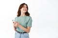 Young modern girl standing with smartphone and look aside at copy space, smiling while chatting on social media app Royalty Free Stock Photo