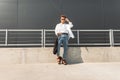 Young modern hipster man in a white shirt in blue jeans in sunglasses with a black bag in red sandals is standing in the city Royalty Free Stock Photo