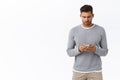 Young modern man in grey sweater using smartphone with busy serious expression, wear wireless earphones, holding mobile