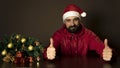 Young modern santa claus is sitting at a table near the tree. Emotional portrait. Thumds up
