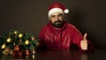 Young modern santa claus is sitting at a table near the tree. Emotional portrait. Thumds up