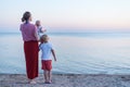 Young mother with baby and eldest son stand on seaside. Rear view. Family on beach at sunset