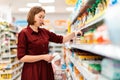 A young mother chooses baby food on a supermarket shelf. In the background, in a blur, a toddler sits in a grocery cart. The