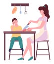 Young mother cooking with her son, cutting tomato at kitchen, people sitting near kitchen table