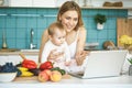 Young mother is cooking and playing with her baby daughter in a modern kitchen. Healthy food concept. Looking at laptop, Royalty Free Stock Photo