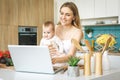 Young mother is cooking and playing with her baby daughter in a modern kitchen setting. Healthy food concept. Looking at laptop, Royalty Free Stock Photo