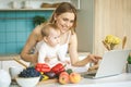 Young mother is cooking and playing with her baby daughter in a modern kitchen setting. Healthy food concept. Looking at laptop, Royalty Free Stock Photo