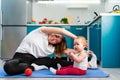 A young mother is doing yoga on a mat in the kitchen, and her baby is sitting next to her. The concept of home sports training Royalty Free Stock Photo