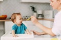 A young mother feeds her young son, who is sitting in a high baby feeding chair. Care, motherhood, childhood Royalty Free Stock Photo