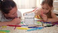 Young mother and her beautiful daughter, paint a paper house, lying on the floor at home, lifestyle, creativity