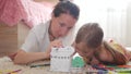 Young mother and her beautiful daughter, paint a paper house, lying on the floor at home, lifestyle, creativity