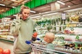 Young mother with her cute baby in shopping trolley walks around supermarket and talks on the phone. Beautiful mom and Royalty Free Stock Photo