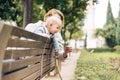 Young mother with her cute infant baby boy child leaning over back of wooden bench towards bushes in city park, holding Royalty Free Stock Photo