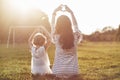 Young mother and her daughter sitting on the grass and making heart symbol to the sun Royalty Free Stock Photo