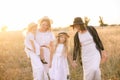 A young mother with her daughters and an aunt with blond hair in white dresses at sunset in the summer in a field of countryside.