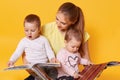 Young mother and her little babies daughters reading books, look on colourful pages, momny keeps children in her knees while Royalty Free Stock Photo