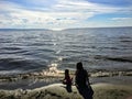 A young mother and her toddler daughter sitting together alone on a sandy beach watching the sparkling lake water