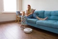 A young mother with her toddler daughter watching a modern robot vacuum cleaner work on the floor in the living room