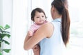 Young mother holding her smiling cute little Caucasian 7 months newborn baby on her hands, standing near window at home. Happy