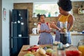 Young mother in the home kitchen with her little daughter, cutting up vegetables and tasting cucumber slices, girl acting silly