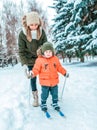 A young mother insures a child from back on children skis young son of 3-5 years old, in winter in city against backdrop