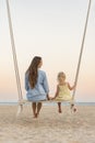 Young mother and little fair-haired girl sit on swing on sea background during sunset. Mom and daughter on the beach over yellow Royalty Free Stock Photo