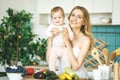 Young mother looking at camera and smiling, cooking and playing with her baby daughter in a modern kitchen setting. Healthy food