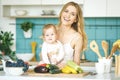 Young mother looking at camera and smiling, cooking and playing with her baby daughter in a kitchen setting. Healthy food