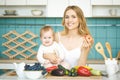 Young mother looking at camera and smiling, cooking and playing with her baby daughter in a modern kitchen setting. Healthy food