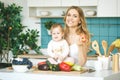 Young mother looking at camera and smiling, cooking and playing with her baby daughter in a modern kitchen setting. Healthy food Royalty Free Stock Photo