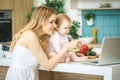 Young mother looking at camera and smiling, cooking and playing with her baby daughter in a modern kitchen. Using laptop. Healthy Royalty Free Stock Photo