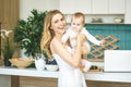 Young mother looking at camera and smiling, cooking and playing with her baby daughter in a modern kitchen. Using phone. Healthy Royalty Free Stock Photo