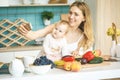 Young mother looking at camera and smiling, cooking and playing with her baby daughter in a modern kitchen. Using phone, making Royalty Free Stock Photo