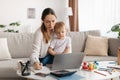 Young mother multitasking, working remotely on laptop while taking care of kid son, sitting on sofa with child on hands