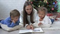 Young mother other with two kids reading a book on the floor near Christmas tree