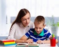 Young mother sitting at a table at home helping her small son with his homework from school as he writes notes in a notebook Royalty Free Stock Photo