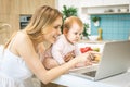 Young mother smiling, cooking and playing with her baby daughter in a kitchen. Using laptop. Healthy food concept. Working Royalty Free Stock Photo