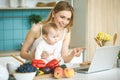 Young mother smiling, cooking and playing with her baby daughter in a modern kitchen. Using laptop. Healthy food concept. Royalty Free Stock Photo