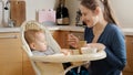 Young mother smiling at her baby son sitting in highchair at kitchen. Concept of parenting, healthy nutrition and baby care Royalty Free Stock Photo