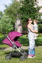 Young mother in summer light dress holding her toddler daughter in arms, woman standing close to baby stroller on green lawn Royalty Free Stock Photo