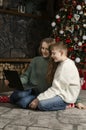 Young mother and teenage son sit near Christmas tree with laptop. Older sister and brother talk to their parents with video call Royalty Free Stock Photo