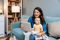 Young mother trying to entertain her newborn baby by playing cartoons and funny videos on digital tablet while sitting on sofa at Royalty Free Stock Photo