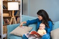 Young mother trying to entertain her newborn baby by reading a fairytale books while sitting on sofa at home. Woman read a book